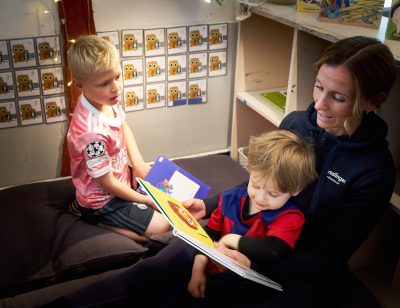 pedagog sitter med ett barn i knät och läser bok medan ett annat barn tittar på
