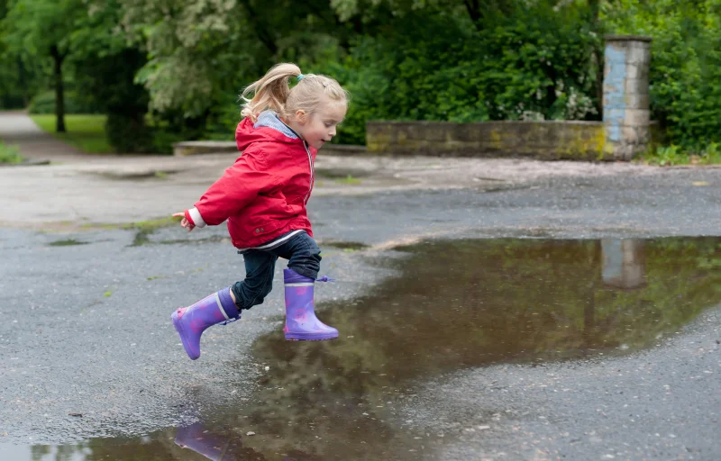 Flicka med funktionella kläder och gummistövlar hoppar i vattenpöl