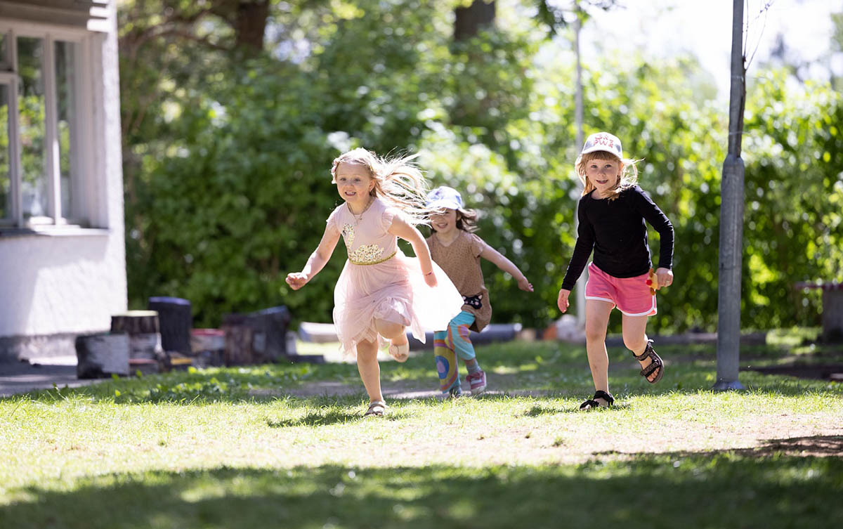 Tre glada flickor i sommarkläder springer på en grön gräsmatta