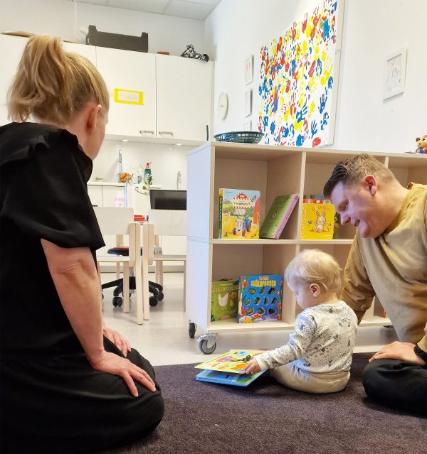 Ett barn och en man sitter på golvet och läser en bok på Minipyss medan en kvinna ser på